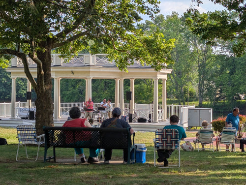 Musicians performing at a white park pavilion with an audience seated beneath leafy trees.